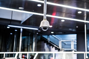 A surveillance camera is mounted on the ceiling in a modern, metallic interior environment. The ceiling features multiple recessed lights and linear designs, contributing to a sleek and industrial atmosphere. In the background, a staircase with metal railings is visible, indicating a public or corporate setting.