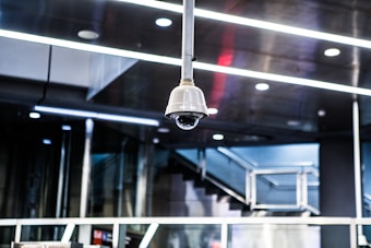 A surveillance camera is mounted on the ceiling in a modern, metallic interior environment. The ceiling features multiple recessed lights and linear designs, contributing to a sleek and industrial atmosphere. In the background, a staircase with metal railings is visible, indicating a public or corporate setting.