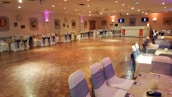A large banquet hall features a polished wooden floor surrounded by elegantly draped chairs with dark sashes. Soft, ambient lighting from wall-mounted fixtures and small ceiling lights creates a warm and inviting atmosphere. The room is decorated with framed pictures on the walls, and a small bar area is visible in the background. Tables are set up around the perimeter, enhancing the anticipation of an upcoming event.