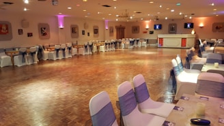 A large banquet hall features a polished wooden floor surrounded by elegantly draped chairs with dark sashes. Soft, ambient lighting from wall-mounted fixtures and small ceiling lights creates a warm and inviting atmosphere. The room is decorated with framed pictures on the walls, and a small bar area is visible in the background. Tables are set up around the perimeter, enhancing the anticipation of an upcoming event.
