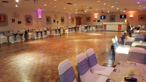 A large banquet hall features a polished wooden floor surrounded by elegantly draped chairs with dark sashes. Soft, ambient lighting from wall-mounted fixtures and small ceiling lights creates a warm and inviting atmosphere. The room is decorated with framed pictures on the walls, and a small bar area is visible in the background. Tables are set up around the perimeter, enhancing the anticipation of an upcoming event.