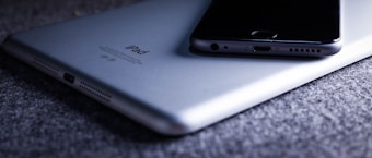 A close-up view of an iPhone placed on top of an iPad on a textured gray surface. Both devices are positioned to reveal their edges and ports, with the iPhone resting diagonally on the iPad.