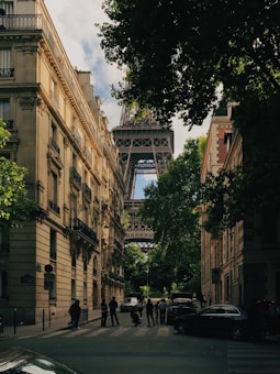 A narrow Parisian street lined with classic French architecture leads to the iconic Eiffel Tower, partially obscured by lush green trees. A group of people is visible on the street, and cars are parked along the sides.