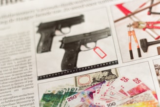 A newspaper page featuring images of two pistols, a set of tools including pliers and a hammer, and various banknotes. The pistols have tags attached, and there is a measuring scale beneath them. The tools appear organized on a separate section, while the banknotes are displayed in denominations of varying values, including bills with colorful patterns and a portrait.