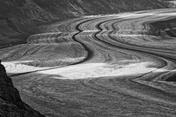 An expansive glacial landscape characterized by curved, flowing lines that suggest movement. The surface of the glacier displays intricate patterns formed by the natural flow of ice, bordered by dark mountain slopes.