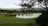 A serene golf course landscape featuring a calm lake surrounded by lush greenery and distant mountains. Trees provide shade in the foreground, while the course features well-manicured grass and sand bunkers. The overall scene is peaceful and well-maintained.