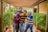Four friends are standing close together in a covered outdoor walkway. They are smiling and appear happy, with one person holding a camera. The setting is lush with greenery, and a wooden pergola-like structure runs overhead.