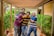 Four friends are standing close together in a covered outdoor walkway. They are smiling and appear happy, with one person holding a camera. The setting is lush with greenery, and a wooden pergola-like structure runs overhead.