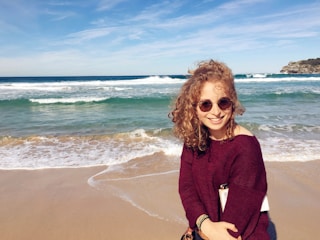 A woman with curly hair is smiling at the camera while standing on a sandy beach. She is wearing sunglasses and a burgundy sweater. Behind her, there are gentle waves in the ocean under a partly cloudy sky.