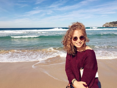 A woman with curly hair is smiling at the camera while standing on a sandy beach. She is wearing sunglasses and a burgundy sweater. Behind her, there are gentle waves in the ocean under a partly cloudy sky.