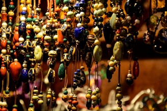 A collection of colorful, intricately designed beads hanging closely together, featuring a variety of shapes and hues, including blues, greens, oranges, and yellows. The beads are strung on thin cords and are densely arranged, creating a rich texture and vibrant visual effect against a soft-focus background.