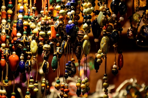 A collection of colorful, intricately designed beads hanging closely together, featuring a variety of shapes and hues, including blues, greens, oranges, and yellows. The beads are strung on thin cords and are densely arranged, creating a rich texture and vibrant visual effect against a soft-focus background.