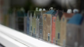 A row of small, colorful model houses with various pastel shades arranged in a line, creating a miniature street scene. The focus is on the intricate details of the houses, such as the windows and rooftops, with a slight blur in the background and foreground giving a sense of depth.