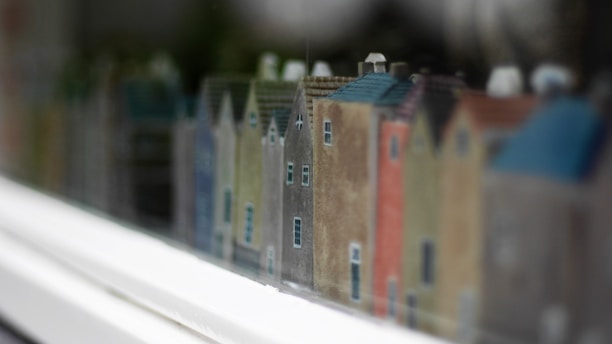 A row of small, colorful model houses with various pastel shades arranged in a line, creating a miniature street scene. The focus is on the intricate details of the houses, such as the windows and rooftops, with a slight blur in the background and foreground giving a sense of depth.