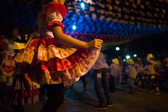 A festive dance scene featuring people in traditional attire, with a focus on a person wearing a vibrant, layered dress with shades of red and pink. The atmosphere is lively with decorations overhead and people in cowboy hats dancing in the background.