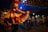 A festive dance scene featuring people in traditional attire, with a focus on a person wearing a vibrant, layered dress with shades of red and pink. The atmosphere is lively with decorations overhead and people in cowboy hats dancing in the background.