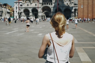 A person with blonde hair, wearing a white top, is walking through a bustling square towards a large, ornate building with arches and intricate architectural features. The area is populated with many people, suggesting a tourist destination.