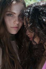Two individuals with long, wavy hair are close together, with one partly leaning on the other. Both have serious expressions. Natural light creates soft shadows on their faces, and the outdoor setting gives a gentle, organic feel to the scene.