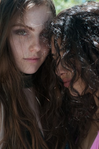 Two individuals with long, wavy hair are close together, with one partly leaning on the other. Both have serious expressions. Natural light creates soft shadows on their faces, and the outdoor setting gives a gentle, organic feel to the scene.