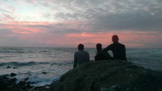 three men sitting on gray rock in front of body of water in Pointe de la Torche France