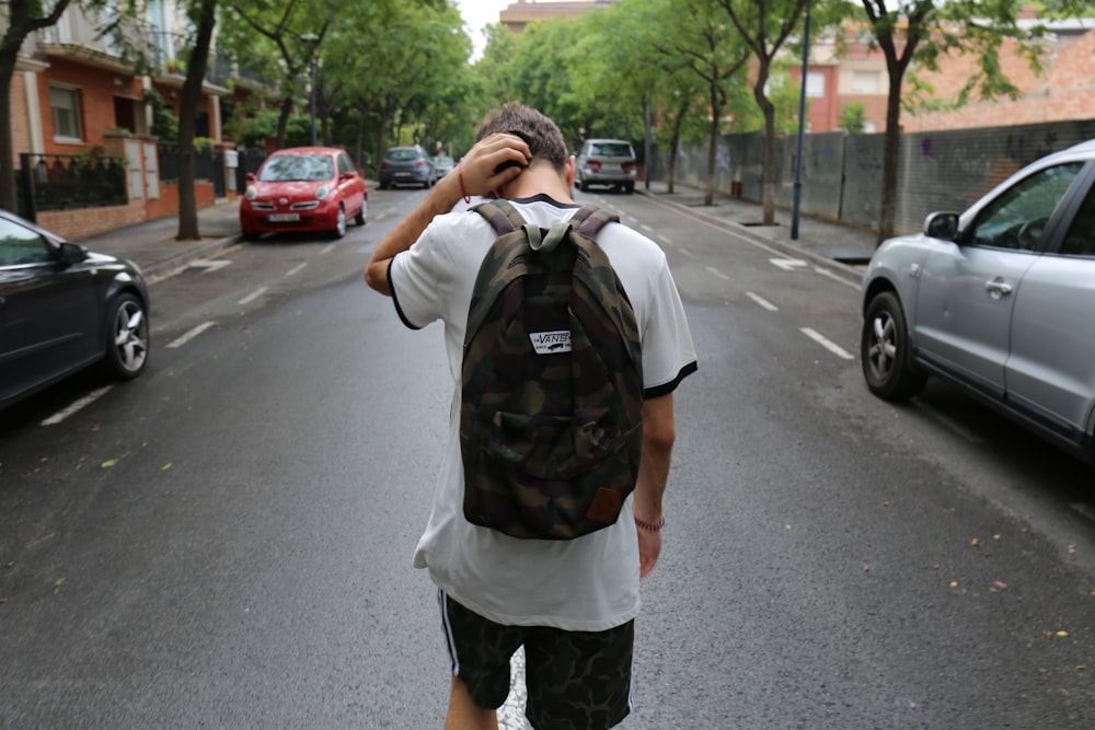 ragazzo che indossa camicia bianca e pantaloncini neri che trasporta zaino in piedi sulla strada di cemento nero tra veicoli e alberi durante il giorno