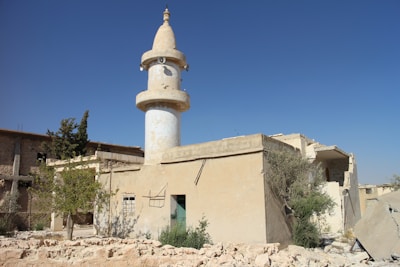 A weathered beige building with a tall cylindrical minaret, partially damaged, sits under a clear blue sky. Vegetation, including small trees and shrubs, grows around and inside the structure, which is surrounded by rubble and debris. The building shows signs of neglect and destruction.