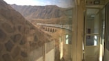 A scenic railway journey showcases a train crossing a viaduct in a mountainous region. The view from inside the train features reflections on the glass and an open door leading to an empty carriage interior, with rugged hills and clear skies in the background.