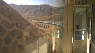 A scenic railway journey showcases a train crossing a viaduct in a mountainous region. The view from inside the train features reflections on the glass and an open door leading to an empty carriage interior, with rugged hills and clear skies in the background.