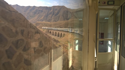 A scenic railway journey showcases a train crossing a viaduct in a mountainous region. The view from inside the train features reflections on the glass and an open door leading to an empty carriage interior, with rugged hills and clear skies in the background.