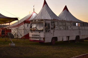 An old, worn-out bus sits next to a large circus tent with red and white stripes. The bus appears to be part of the circus setup, with a weathered and rustic appearance, indicating extensive use. Surrounding the bus and tent is a grassy field, and a satellite dish is visible nearby. The sky is a soft gradient, suggesting twilight or dawn.