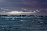 Vast ocean waves under a dramatic sky. Distant shoreline with small houses and vegetation. Clouds appear heavy and layered, indicating possible approaching weather.