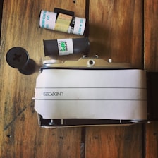 A vintage film camera rests on a wooden surface alongside a roll of film and a film canister. The camera is partially wrapped in a white paper marked 'UNEXPOSED'. The wooden background adds a rustic feel to the scene.