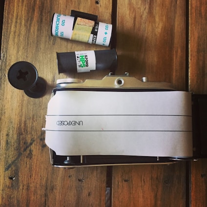 A vintage film camera rests on a wooden surface alongside a roll of film and a film canister. The camera is partially wrapped in a white paper marked 'UNEXPOSED'. The wooden background adds a rustic feel to the scene.