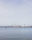 A harbor scene with a large historical sailing ship with multiple masts anchored alongside, surrounded by several smaller sailboats. The water is calm, and the sky is clear with a pale blue hue. In the background, several modern vessels are docked along a breakwater.