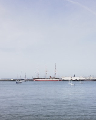 A harbor scene with a large historical sailing ship with multiple masts anchored alongside, surrounded by several smaller sailboats. The water is calm, and the sky is clear with a pale blue hue. In the background, several modern vessels are docked along a breakwater.