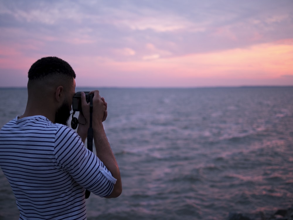 A man in a striped shirt taking a picture of the ocean.
