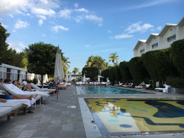 A luxurious outdoor pool area surrounded by lounge chairs and umbrellas, with people relaxing under a clear blue sky. The pool is flanked by tall, trimmed hedges and modern resort buildings. Palm trees are visible in the background, enhancing the tropical ambiance.
