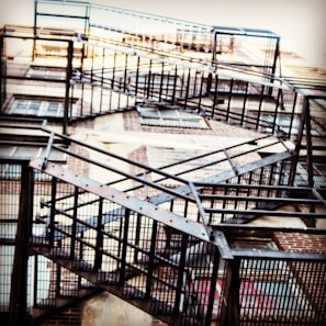 A series of metal fire escape stairs ascend along the side of a brick building. The structure features a crisscross pattern and is composed of dark metal, creating a geometric and industrial look.