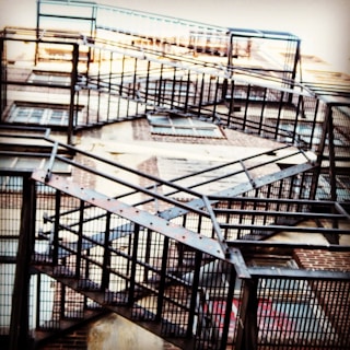 A series of metal fire escape stairs ascend along the side of a brick building. The structure features a crisscross pattern and is composed of dark metal, creating a geometric and industrial look.