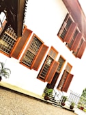 The image features a two-story building characterized by multiple windows with red wooden shutters. The white walls of the building contrast with the red accents of the window shutters and borders. Potted plants are placed near the steps leading to a black iron gate at the entrance. The ground is covered with gravel.
