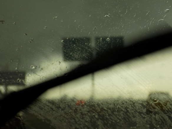 Raindrops cover a windshield as a dark, stormy sky looms in the background. The view is partially obscured by a windshield wiper, with faint images of road signs visible in the distance.
