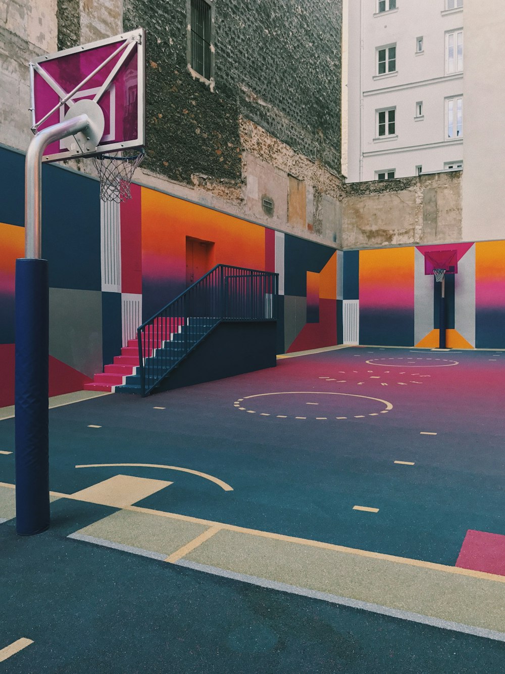 昼間はコンクリートの建物の横の黒、紫、オレンジのバスケットボールコート