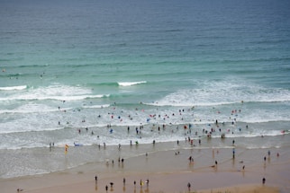 A busy beach scene with numerous people enjoying the ocean, some swimming and others surfing. The beach is lined with individuals in wetsuits and swimsuits, sharing the waves with various surfboards. The water ranges from light to deep blue, with gentle waves rolling in.