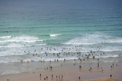 A busy beach scene with numerous people enjoying the ocean, some swimming and others surfing. The beach is lined with individuals in wetsuits and swimsuits, sharing the waves with various surfboards. The water ranges from light to deep blue, with gentle waves rolling in.