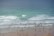 A busy beach scene with numerous people enjoying the ocean, some swimming and others surfing. The beach is lined with individuals in wetsuits and swimsuits, sharing the waves with various surfboards. The water ranges from light to deep blue, with gentle waves rolling in.