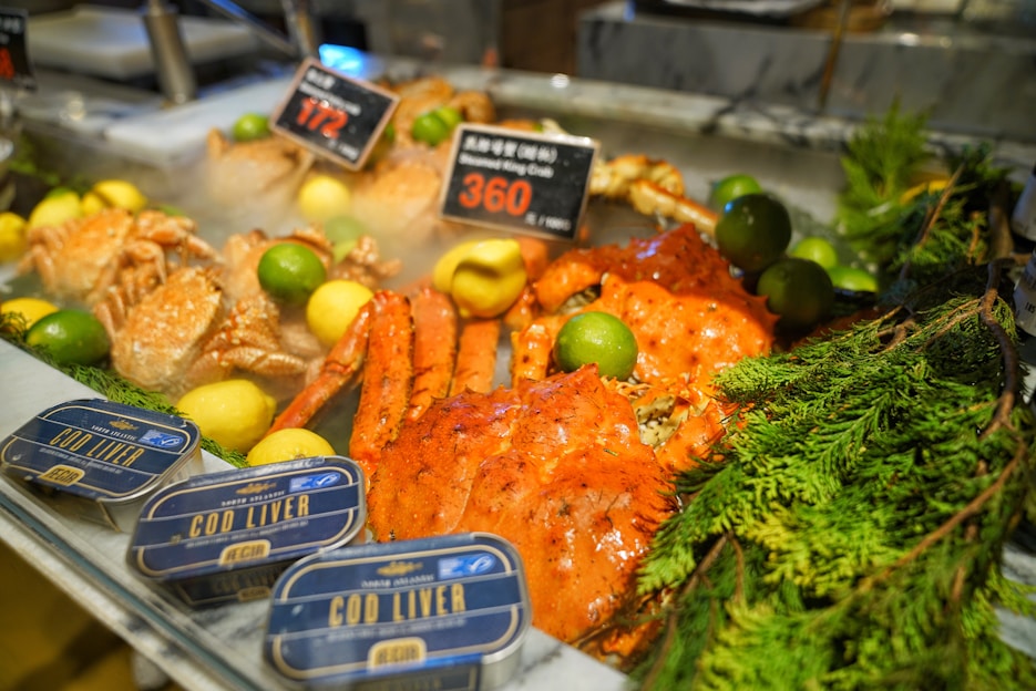 An assortment of seafood is displayed on a chilled counter, featuring bright red crab legs and whole crabs, surrounded by lemons and limes. Tins of cod liver are neatly arranged in the foreground with sprigs of green foliage adding a touch of freshness. Price tags indicate the cost of the items, with steam subtly rising from the cold display.