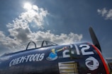 A vintage aircraft with a dark blue fuselage is parked under a bright sky with scattered clouds. The side of the plane features painted artwork and bold white numbers and letters, '212' and 'CHEM-TOOLS'. The artwork depicts a cartoon figure in blue overalls holding a tool. The aircraft canopy is partially visible, and the sunlight creates a dramatic contrast with the clouds.