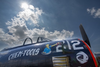 A vintage aircraft with a dark blue fuselage is parked under a bright sky with scattered clouds. The side of the plane features painted artwork and bold white numbers and letters, '212' and 'CHEM-TOOLS'. The artwork depicts a cartoon figure in blue overalls holding a tool. The aircraft canopy is partially visible, and the sunlight creates a dramatic contrast with the clouds.