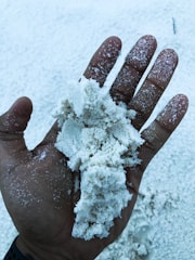A hand holding a pile of fine, white sand. The sand is scattered across the palm and fingers, with more sand visible in the background. The hand is dark-skinned, and the contrast highlights the texture of the sand.