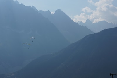 Two paragliders soar through a misty mountain landscape with rugged peaks and scattered clouds. The scene portrays a sense of adventure and tranquility against a backdrop of towering mountains.